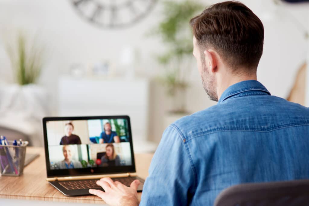 Mann schaut auf seinen Laptop, auf dem mehrere Personen zu sehen sind, die alle gemeinsam in einer Video Konferenz im Homeoffice sind