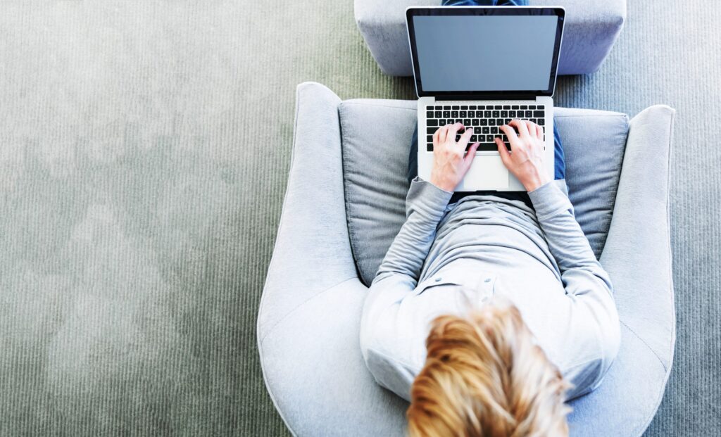 Eine Person sitzt auf einem Sessel und hat die Hände auf der Tastatur des Laptops liegen, der auf dem Schoß der Person ist.